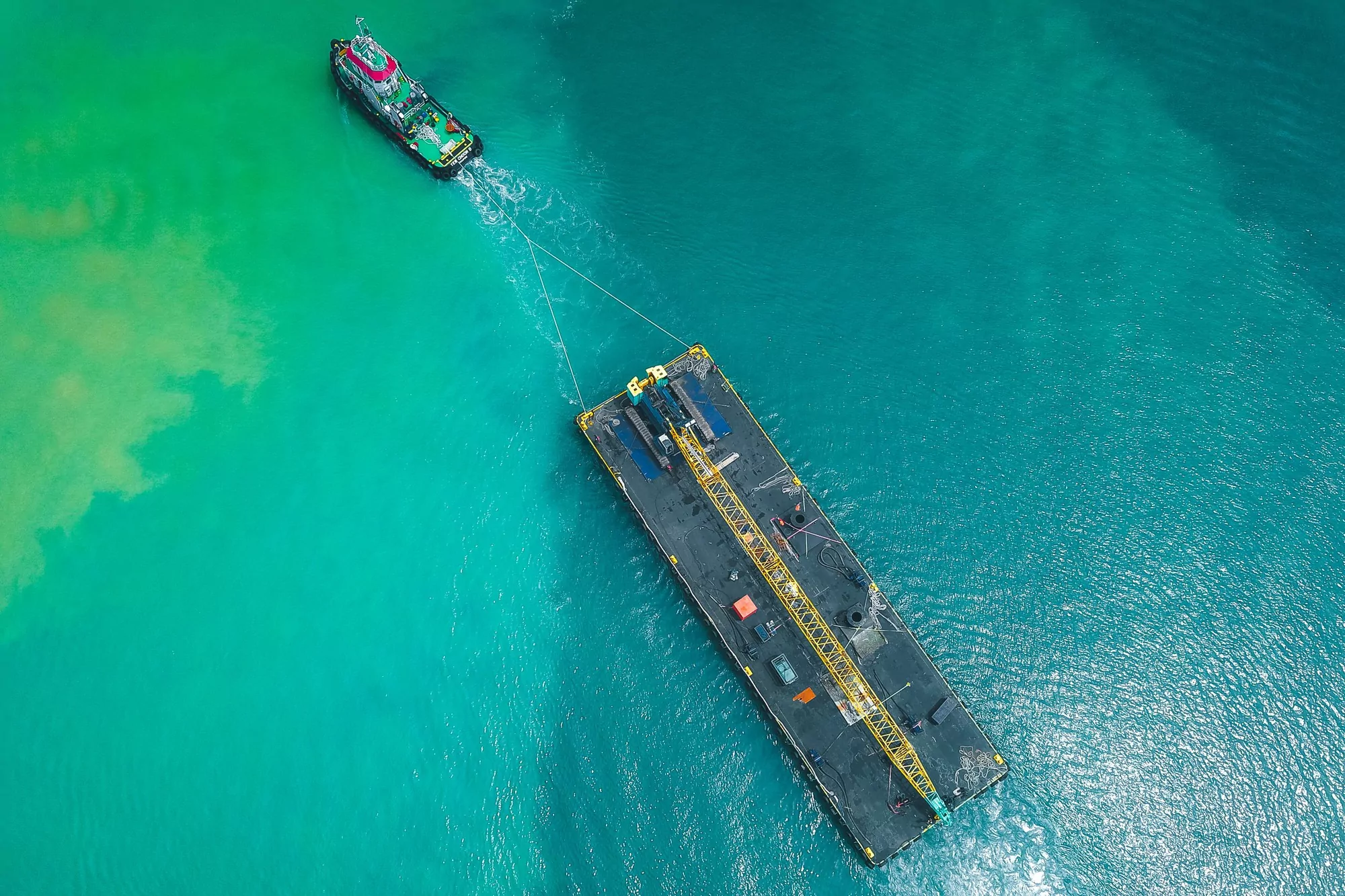 Tug towing a heavy barge across shallow turquoise water seen from the air